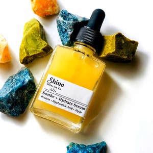 May include: A clear glass bottle with a dropper filled with a yellow serum. The bottle is labeled "Shine Skincare Co. Soothe + Hydrate Serum Protein - Hyaluronic Acid - Algae"