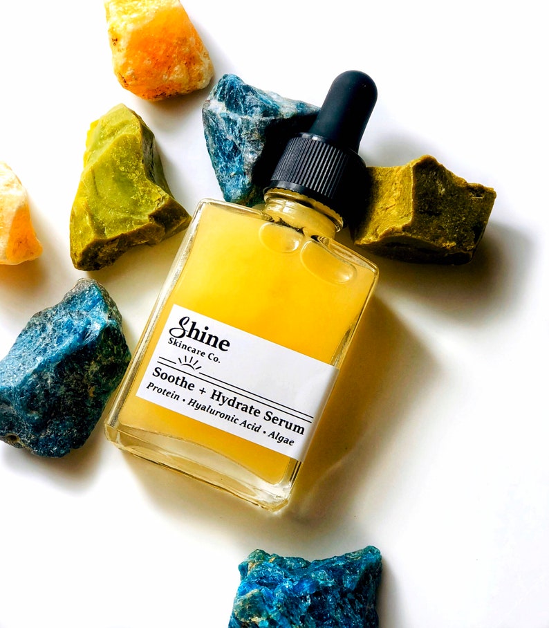 May include: A clear glass bottle with a dropper filled with a yellow serum. The bottle is labeled "Shine Skincare Co. Soothe + Hydrate Serum Protein - Hyaluronic Acid - Algae"