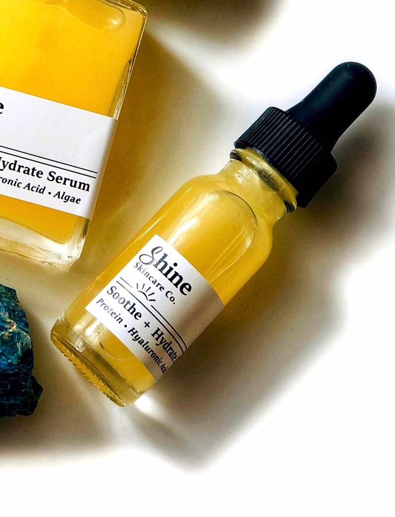 May include: A yellow glass bottle with a black dropper top. The bottle is labeled "Shine Skincare Co." and "Soothe + Hydrate Protein. Hyaluronic Acid."