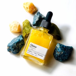 May include: A yellow glass bottle with a black dropper top and a white label that reads "Shine Skincare Co. CoQ10 + Vit C Serum Vit E, CoQ10, Vit C". The bottle is surrounded by blue and green stones.