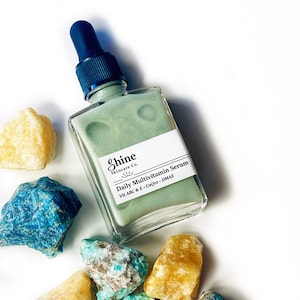 Op de afbeelding: Een groene glazen fles met een zwarte druppelaar. De fles is gelabeld met "Shine Skincare Co. Daily Multivitamin Serum Vit A B C & E, CoQ10, DMAE".