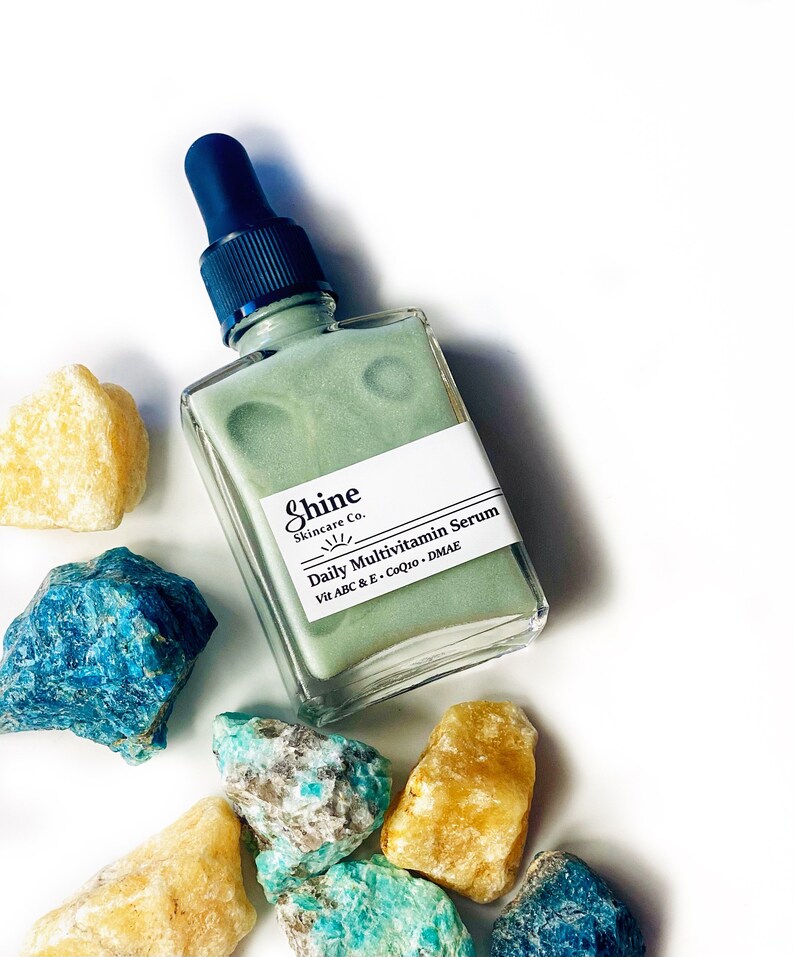 Op de afbeelding: Een groene glazen fles met een zwarte druppelaar. De fles is gelabeld met "Shine Skincare Co. Daily Multivitamin Serum Vit A B C & E, CoQ10, DMAE".