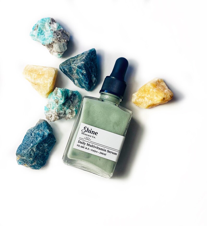 Op de afbeelding: Een groene glazen fles met een druppelaar, met het label "Shine Skincare Co. Daily Multivitamin Serum Vit A, B, C & E - CoQ10 - DMAE" en omringd door kleine blauwe en gele stenen.
