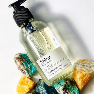 Pu&ograve; includere: Una bottiglia di vetro trasparente con un tappo a pompa nero riempita di un liquido trasparente. La bottiglia ha un'etichetta bianca con il testo "Shine Skincare Co." e "Glycolic Cleanser Burdock, Lemon Peel, Allantoin". La bottiglia &egrave; appoggiata su una superficie bianca con diversi sassi verdi e gialli.