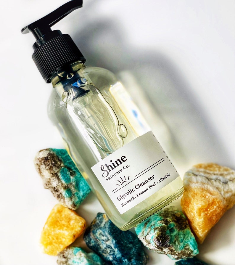 Pu&ograve; includere: Una bottiglia di vetro trasparente con un tappo a pompa nero riempita di un liquido trasparente. La bottiglia ha un'etichetta bianca con il testo "Shine Skincare Co." e "Glycolic Cleanser Burdock, Lemon Peel, Allantoin". La bottiglia &egrave; appoggiata su una superficie bianca con diversi sassi verdi e gialli.