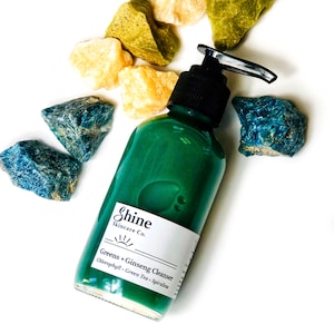Puede incluir: Una botella de vidrio verde con un dispensador de bomba llena de un l&iacute;quido verde. La botella tiene una etiqueta blanca con el texto "Shine Skincare Co. Greens + Ginseng Cleanser Chlorophyll - Green Tea - Spirulina". La botella est&aacute; rodeada de piedras azules y amarillas.