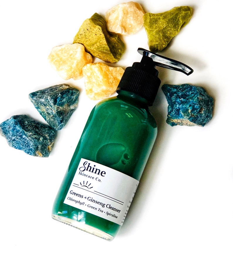 Puede incluir: Una botella de vidrio verde con un dispensador de bomba llena de un l&iacute;quido verde. La botella tiene una etiqueta blanca con el texto "Shine Skincare Co. Greens + Ginseng Cleanser Chlorophyll - Green Tea - Spirulina". La botella est&aacute; rodeada de piedras azules y amarillas.