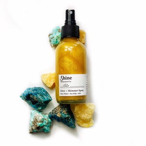 May include: A clear glass bottle of Shine Skincare Co. Glow + Shimmer Spray with a black spray nozzle. The bottle is filled with a golden liquid and surrounded by colorful rocks. The label includes the ingredients: Rose Water, Sea Kelp, and Aloe.