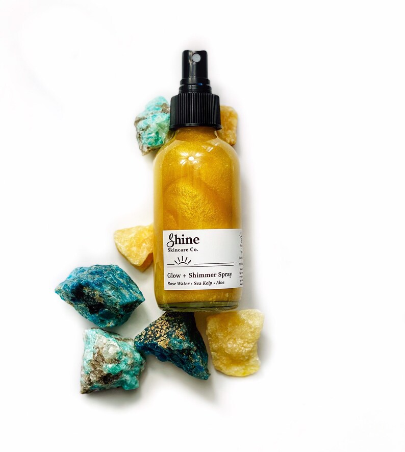 May include: A clear glass bottle of Shine Skincare Co. Glow + Shimmer Spray with a black spray nozzle. The bottle is filled with a golden liquid and surrounded by colorful rocks. The label includes the ingredients: Rose Water, Sea Kelp, and Aloe.