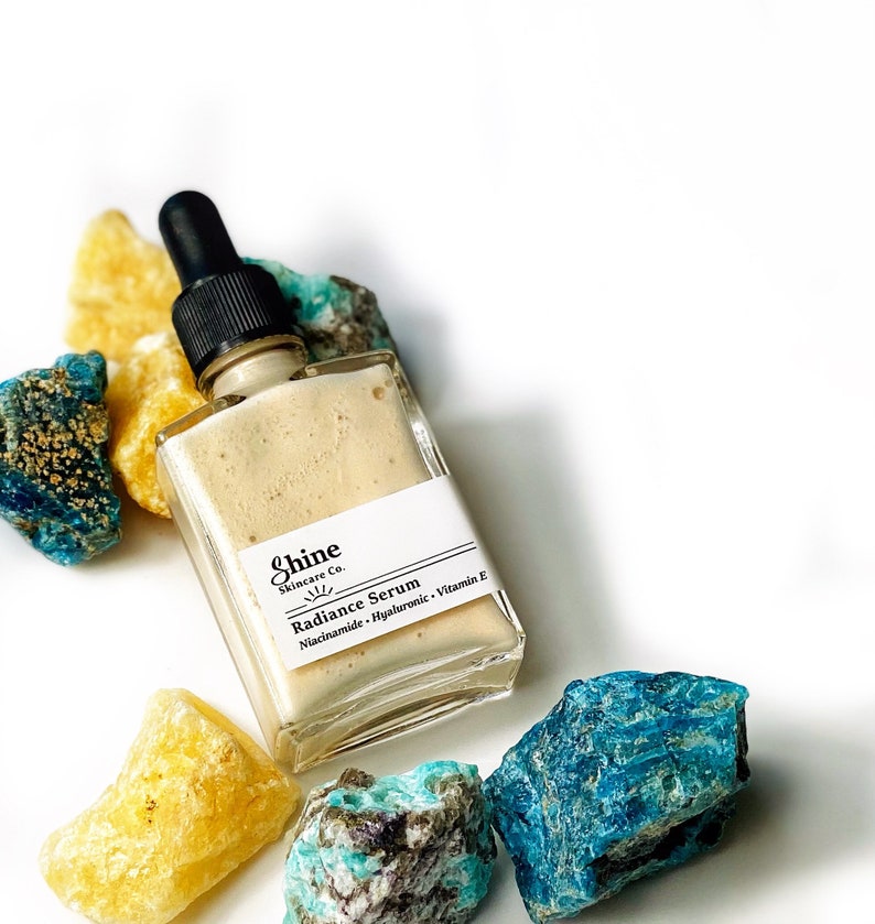 May include: A glass bottle of Shine Skincare Co. Radiance Serum with a dropper top. The bottle is sitting on a white surface with blue and yellow crystals.