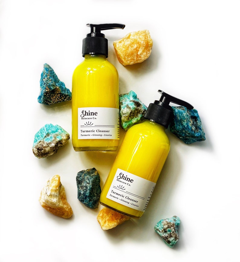 Organic skincare turmeric cleanser in clear glass bottle with pump dispenser. Face wash is bright yellow color and photographed on white background with two bottles. Skincare products surrounded by blue and yellow crystals.