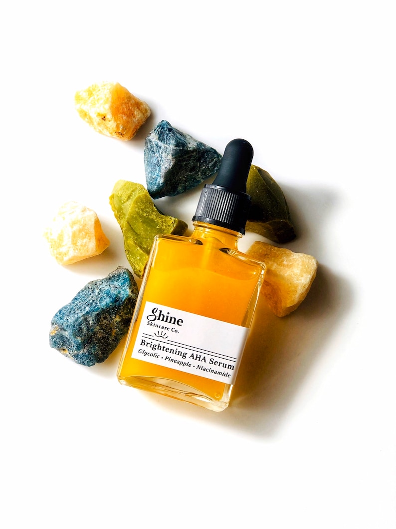 Organic skincare product brightening face serum in clear square glass bottle. Serum is laying on yellow, blue and green crystal gemstones on white background. Shine Skincare label reads brightening aha serum with glycolic, pineapple and niacinamide