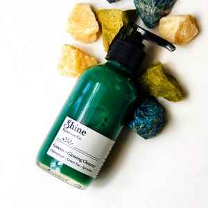 Natural skincare green tea cleanser in clear glass bottle with pump lid. Face wash is a dark green color and thick consistency. White background with blue, green and yellow crystal gemstones around top of bottle.