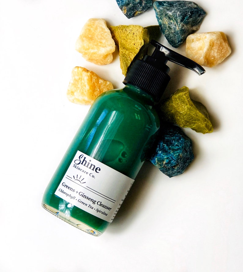 Natural skincare green tea cleanser in clear glass bottle with pump lid. Face wash is a dark green color and thick consistency. White background with blue, green and yellow crystal gemstones around top of bottle.