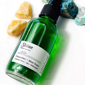 Organic skincare cucumber toner in clear glass bottle with black spray top. Face toner is bright green in color. Photo taken on white background with blue and yellow crystal gemstones along the side of the product.