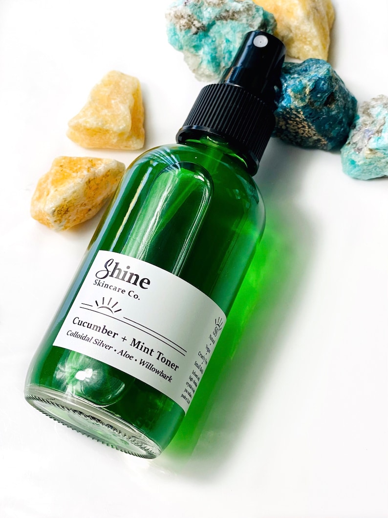 Organic skincare cucumber toner in clear glass bottle with black spray top. Face toner is bright green in color. Photo taken on white background with blue and yellow crystal gemstones along the side of the product.