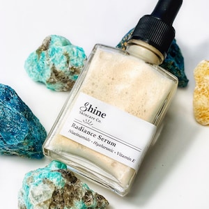 May include: A clear glass bottle with a dropper top filled with a light brown serum. The bottle has a white label with the text "Shine Skincare Co." and "Radiance Serum Niacinamide - Hyaluronic - Vitamin E".