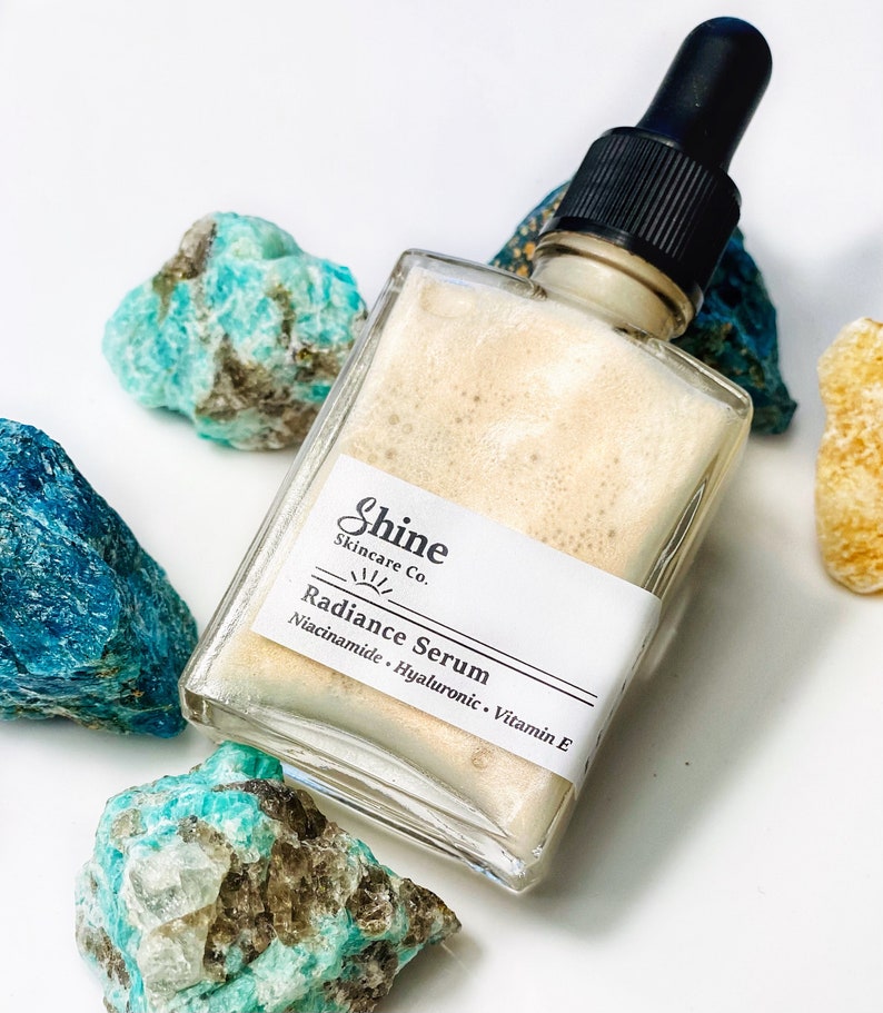May include: A clear glass bottle with a dropper top filled with a light brown serum. The bottle has a white label with the text "Shine Skincare Co." and "Radiance Serum Niacinamide - Hyaluronic - Vitamin E".