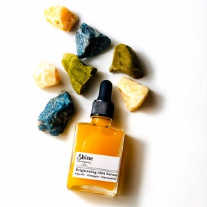 May include: A small amber glass bottle with a black dropper top and a white label that reads "Shine Skincare Co. Brightening AHA Serum Glycolic - Pineapple - Niacinamide". The bottle is surrounded by small yellow, green, and blue stones.