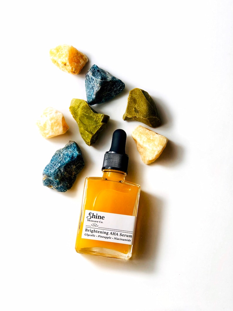 May include: A small amber glass bottle with a black dropper top and a white label that reads "Shine Skincare Co. Brightening AHA Serum Glycolic - Pineapple - Niacinamide". The bottle is surrounded by small yellow, green, and blue stones.