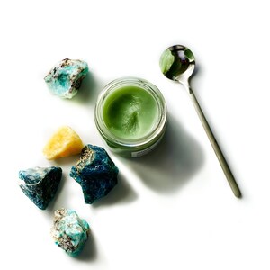 May include: A small jar of green balm with a silver spoon and several blue and green crystals on a white background.