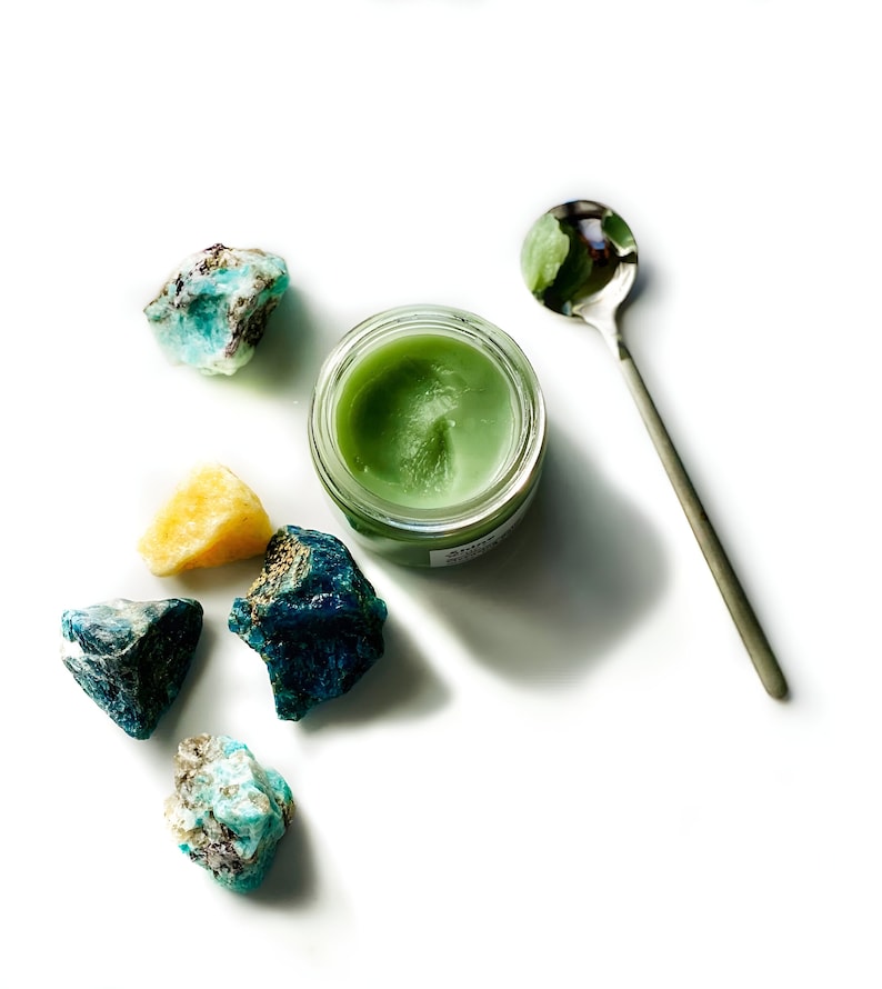 May include: A small jar of green balm with a silver spoon and several blue and green crystals on a white background.