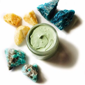 Organic skincare superfood clay face mask is packaged in a clear glass jar. Clay mask is a light green color. Skincare product is photographed showing open top of jar on a white background with yellow and blue crystal gemstones around the face mask.