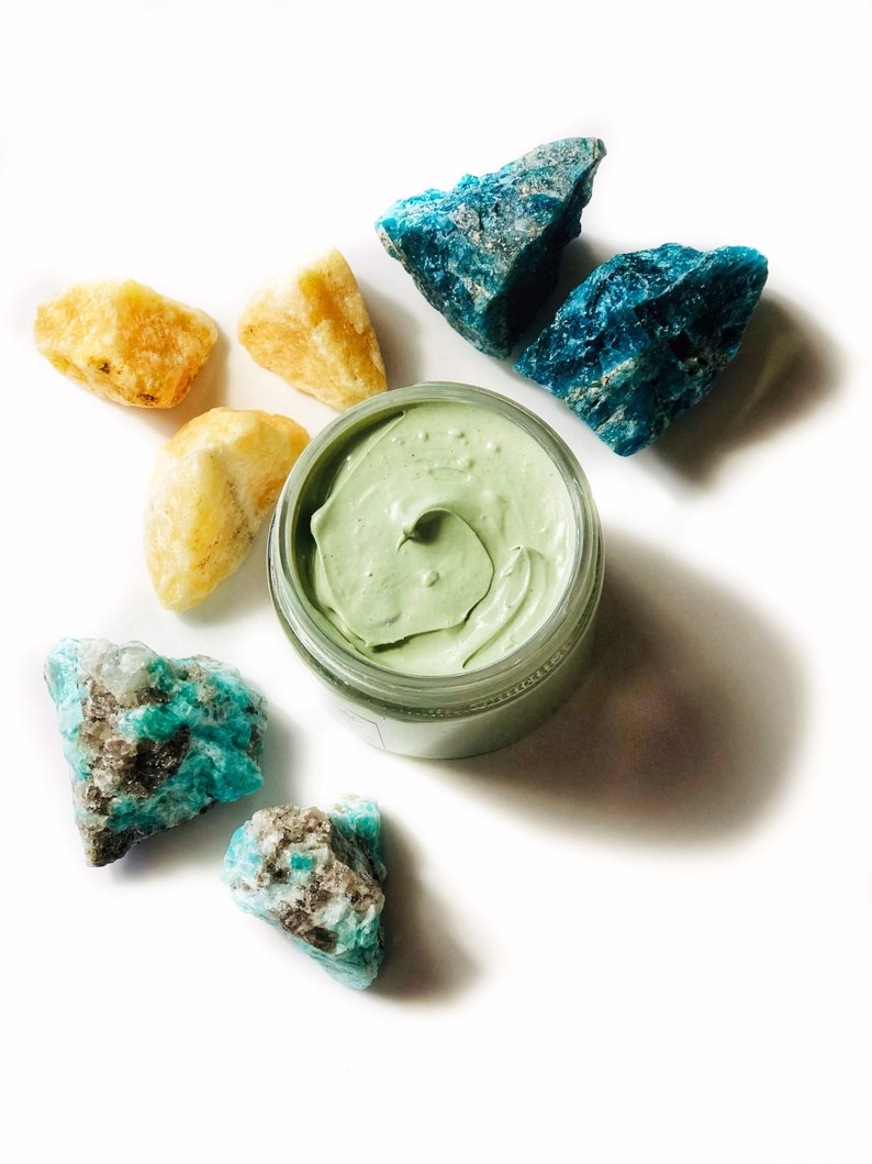 Organic skincare superfood clay face mask is packaged in a clear glass jar. Clay mask is a light green color. Skincare product is photographed showing open top of jar on a white background with yellow and blue crystal gemstones around the face mask.