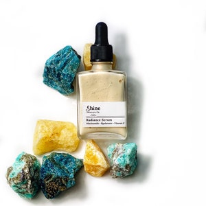 May include: A glass bottle of serum with a dropper top. The bottle is labelled "Shine Skincare Co. Radiance Serum Niacinamide - Hyaluronic - Vitamin E". The bottle is surrounded by yellow and blue gemstones.