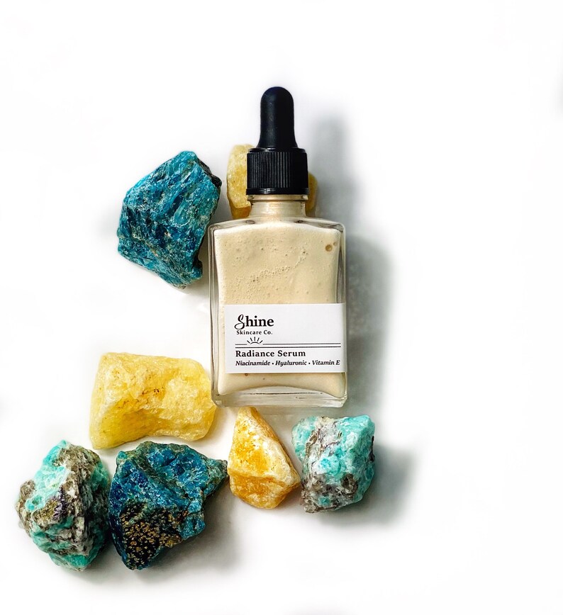 May include: A glass bottle of serum with a dropper top. The bottle is labelled "Shine Skincare Co. Radiance Serum Niacinamide - Hyaluronic - Vitamin E". The bottle is surrounded by yellow and blue gemstones.
