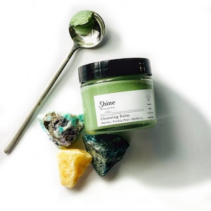 May include: A green cleansing balm jar with a white label that reads "Shine Skincare Co. Cleansing Balm Matcha - Prickly Pear - Mulberry". The jar is sitting on a white surface with three green and yellow crystals.