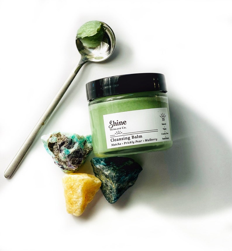 May include: A green cleansing balm jar with a white label that reads "Shine Skincare Co. Cleansing Balm Matcha - Prickly Pear - Mulberry". The jar is sitting on a white surface with three green and yellow crystals.