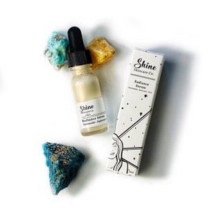 May include: A clear glass bottle with a dropper filled with a light gold liquid. The bottle is labelled "Shine Skincare Co. Radiance Serum Niacinamide - Hyaluronic Acid". The bottle is sitting on a white surface with two blue-green crystals and a yellow crystal.