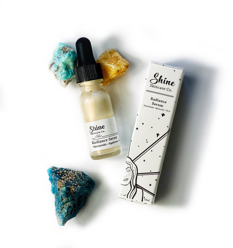 May include: A clear glass bottle with a dropper filled with a light gold liquid. The bottle is labelled "Shine Skincare Co. Radiance Serum Niacinamide - Hyaluronic Acid". The bottle is sitting on a white surface with two blue-green crystals and a yellow crystal.
