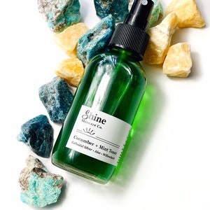 May include: A green glass bottle with a black spray top, labeled "Shine Skincare Co. Cucumber + Mint Toner Colloidal Silver - Aloe - Willowbark". The bottle is surrounded by blue and yellow stones.