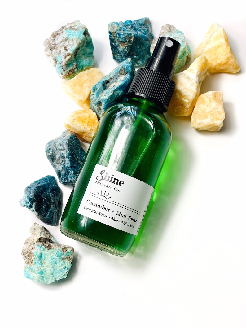 May include: A green glass bottle with a black spray top, labeled "Shine Skincare Co. Cucumber + Mint Toner Colloidal Silver - Aloe - Willowbark". The bottle is surrounded by blue and yellow stones.