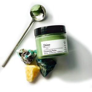 May include: A green glass jar with a black lid containing a green cleansing balm. The jar is labeled "Shine Skincare Co. Cleansing Balm Matcha - Prickly Pear - Mulberry". There are three green, yellow, and blue stones around the jar.