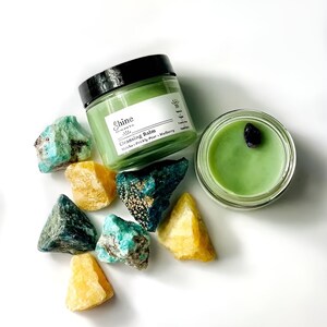 May include: A green glass jar of cleansing balm with the label "Shine Skincare Co. Cleansing Balm Litsea, Prickly Pear, Mulberry" and a green glass jar with a black stone inside. The jars are surrounded by green and yellow crystals.