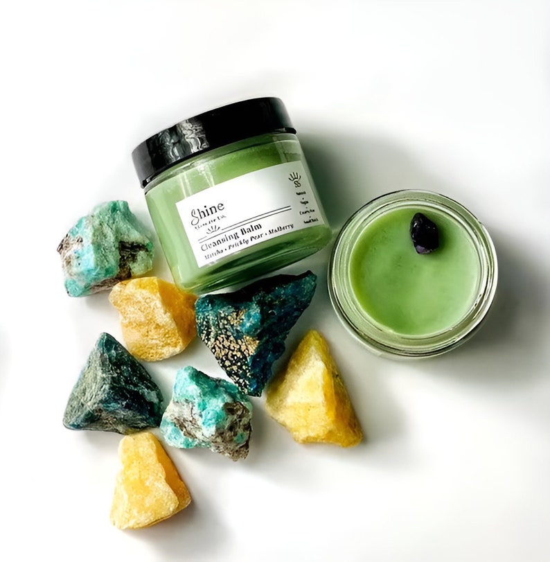 May include: A green glass jar of cleansing balm with the label "Shine Skincare Co. Cleansing Balm Litsea, Prickly Pear, Mulberry" and a green glass jar with a black stone inside. The jars are surrounded by green and yellow crystals.