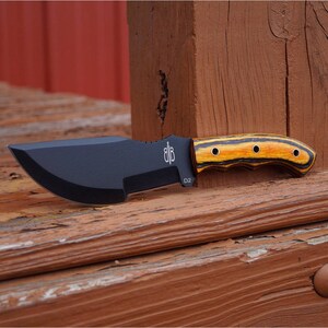 May include: A black and brown hunting knife with a wooden handle. The handle has a black and yellow striped pattern and four brass rivets. The blade is labelled "D2" and has a small logo on the side.