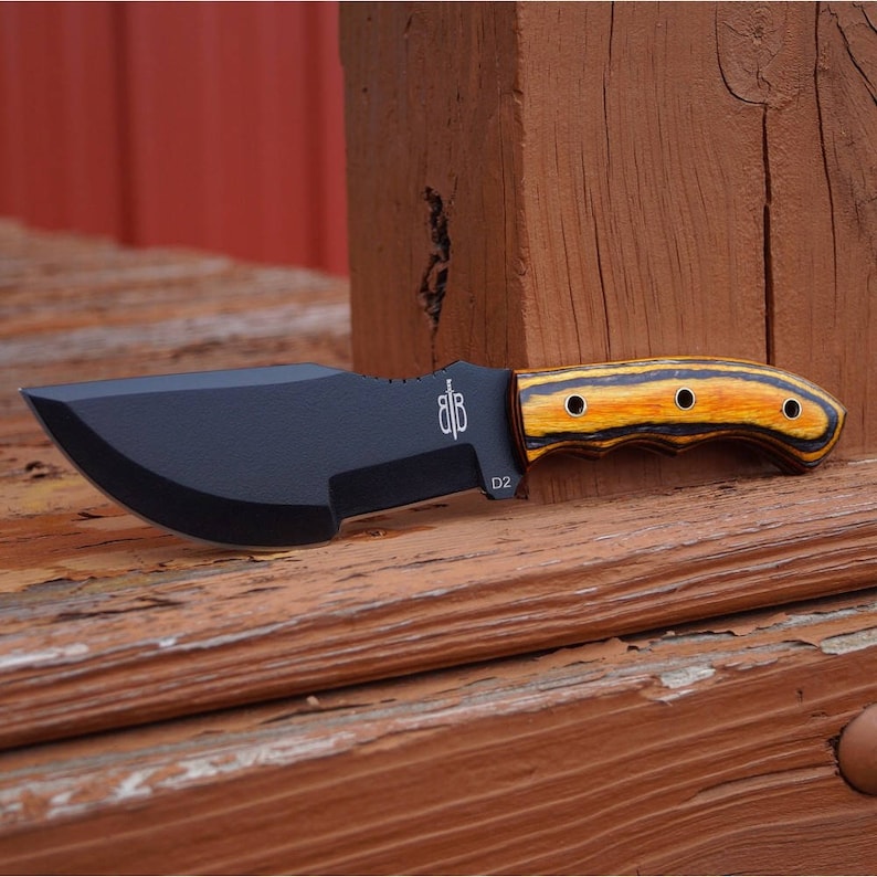 May include: A black and brown hunting knife with a wooden handle. The handle has a black and yellow striped pattern and four brass rivets. The blade is labelled "D2" and has a small logo on the side.