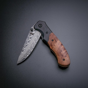 Może przedstawiać: Częściowo otwarty scyzoryk z ostrzem ze stali o wzorze i drewnianą rękojeścią. Rękojeść jest w kolorze jasnobrązowym z ciemnobrązowymi akcentami. Ostrze ma unikalny, wirujący wzór. Nóż jest ustawiony na ciemnym tle.