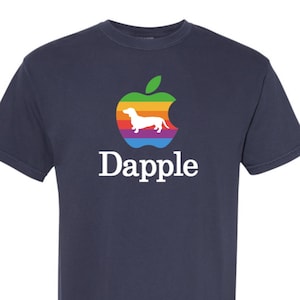 May include: Navy blue t-shirt featuring a colorful apple logo with a dachshund silhouette and the word "Dapple" in white. The apple design incorporates rainbow colors. A casual tee for dog lovers.