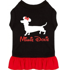 Puede incluir: Vestido negro para perro con una silueta blanca de un dachshund que lleva un lazo rojo con lunares blancos. El texto "Minnie Doxie" est&aacute; impreso en rojo en el vestido.