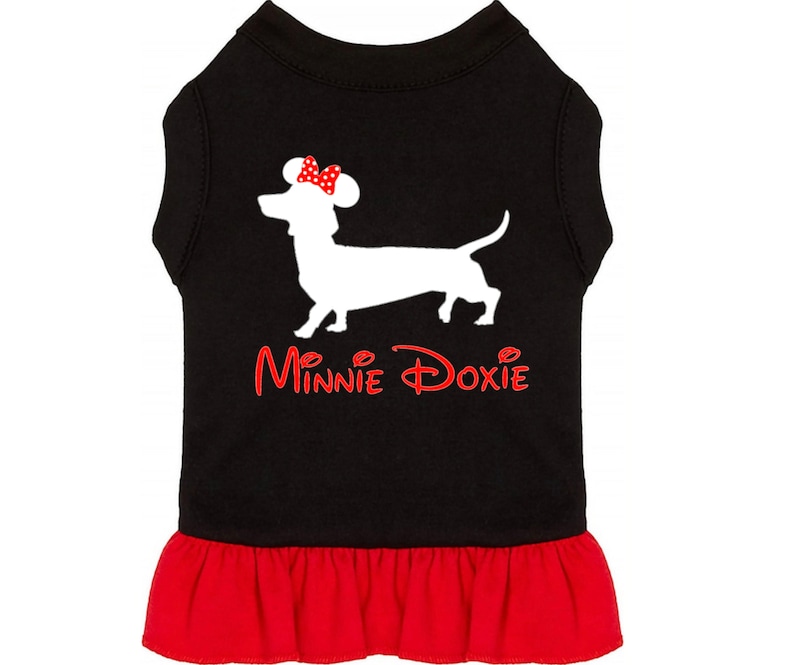 Puede incluir: Vestido negro para perro con una silueta blanca de un dachshund que lleva un lazo rojo con lunares blancos. El texto "Minnie Doxie" est&aacute; impreso en rojo en el vestido.