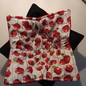 May include: A red and black quilted hot pad with a white background and a pattern of red apples. The hot pad is folded in half and is ready to be used.