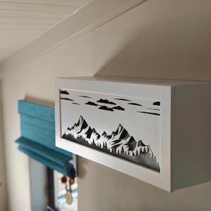May include: A white, rectangular wall-mounted cabinet featuring a black mountain and cloud silhouette design. The cabinet is mounted on a beige wall, with a teal window shade in the background.