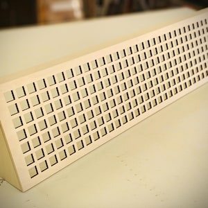 Puede incluir: Una cubierta de ventilación de madera clara con forma rectangular y un patrón de rejilla de cortes cuadrados. La cubierta tiene un lado inclinado y está diseñada para la circulación del aire. La veta de la madera es visible.