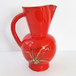 Puede incluir: Una jarra de cerámica de color rojo brillante con acabado esmaltado, que presenta un borde ancho y acampanado junto a un asa curva. El cuerpo de la jarra es redondeado y está decorado con un patrón de estrella en tonos oro, marrón y blanco.