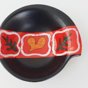 May include: A round black ceramic spoon rest featuring a horizontal red band across the center. The red band contains three white-outlined squares, each displaying a hand-painted design: a green leaf, a yellow heart, and a green leaf.